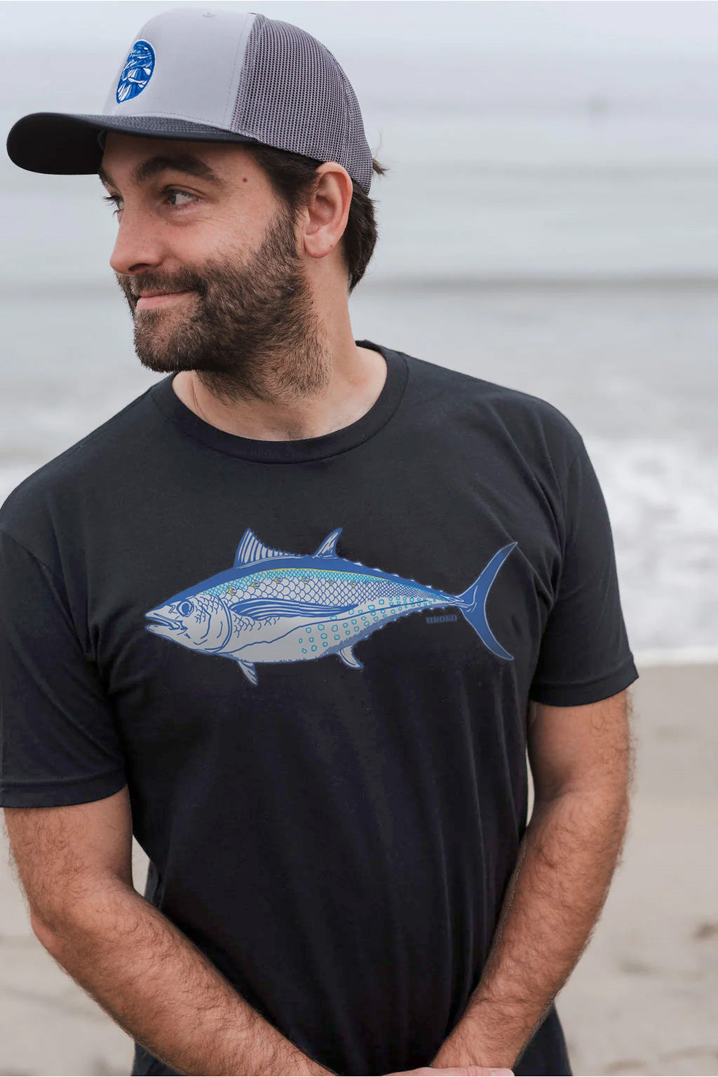 Man wearing a black t-shirt with an albacore tune design, standing on a beach.