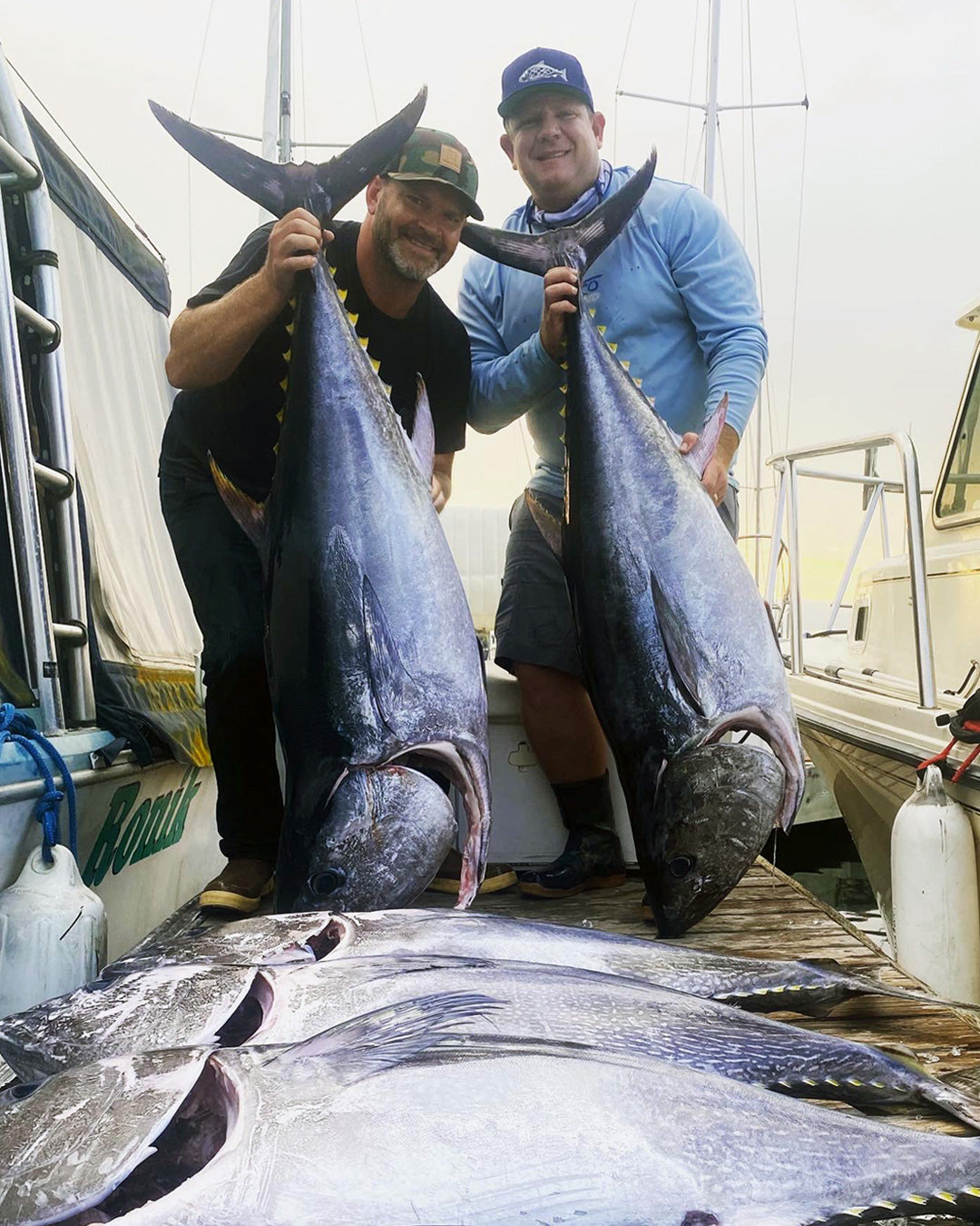 Two men standing on a boat deck holding two large blue tuna fish, with additional tuna laid out on the boat in the foreground.