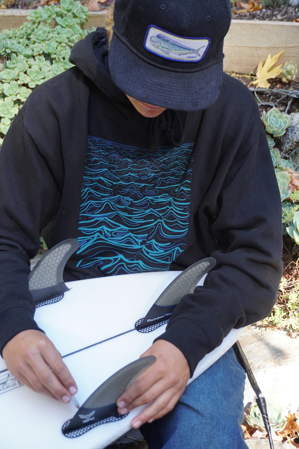 A person outdoors wearing a black hoodie with a blue and black wave graphic, a black cap, and blue jeans, holding a white surfboard with black fins.