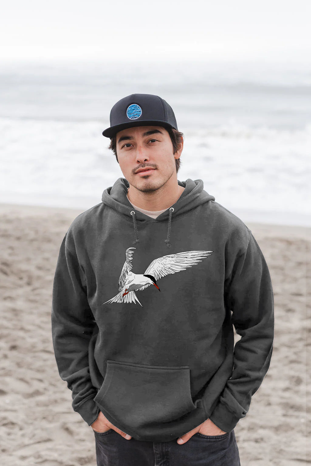 Man wearing a dark gray hoodie with a artic tern design on a beach