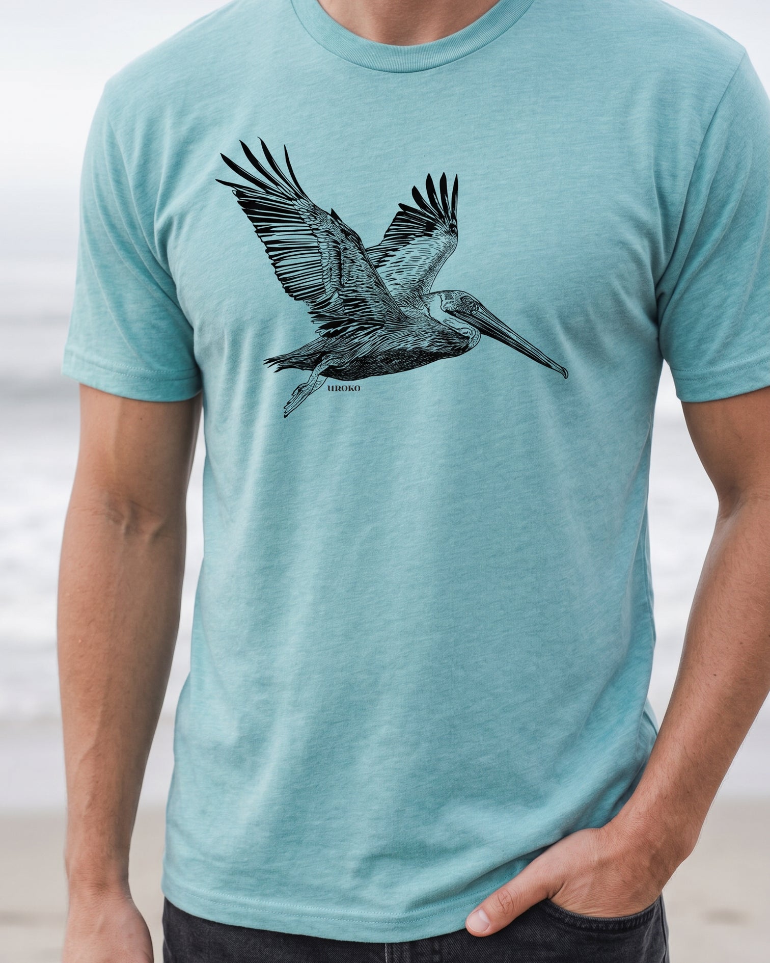 Person wearing a teal t-shirt with a pelican design on a beach.
