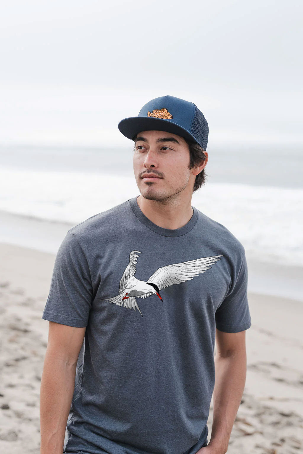 Man wearing a cap and t-shirt with a tern bird design on a beach