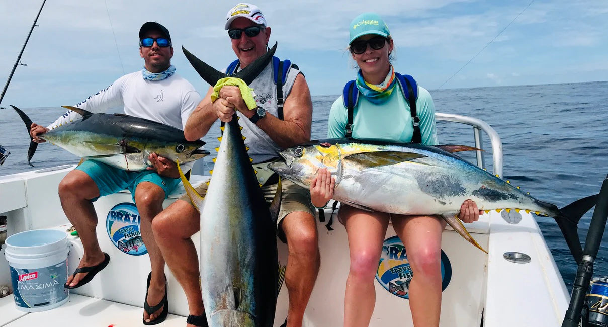 Three people on a boat holding large fish with a clear sky and ocean background