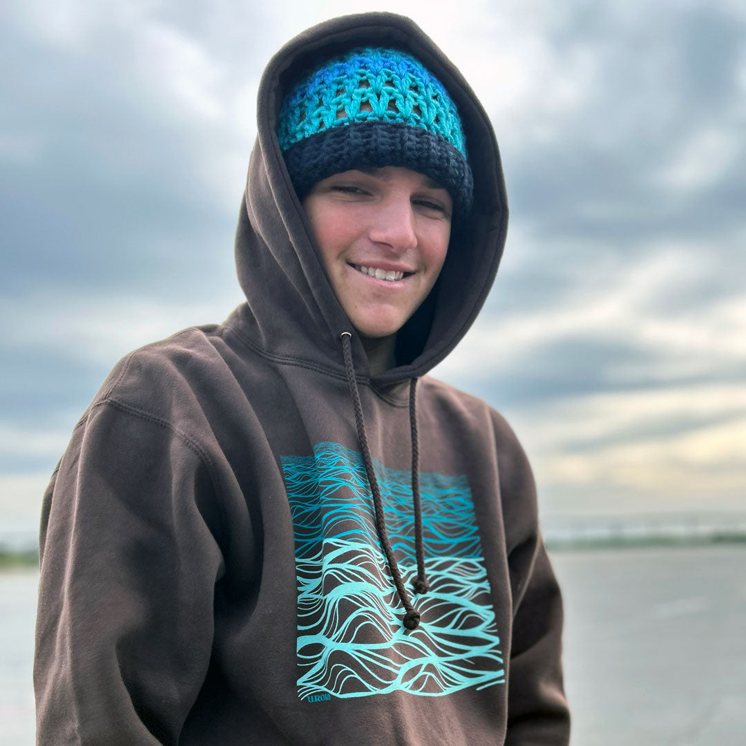 A smiling man outdoors wearing a brown hoodie with a turquoise graphic print on the front and a matching turquoise and dark blue knit beanie, against a cloudy sky background.