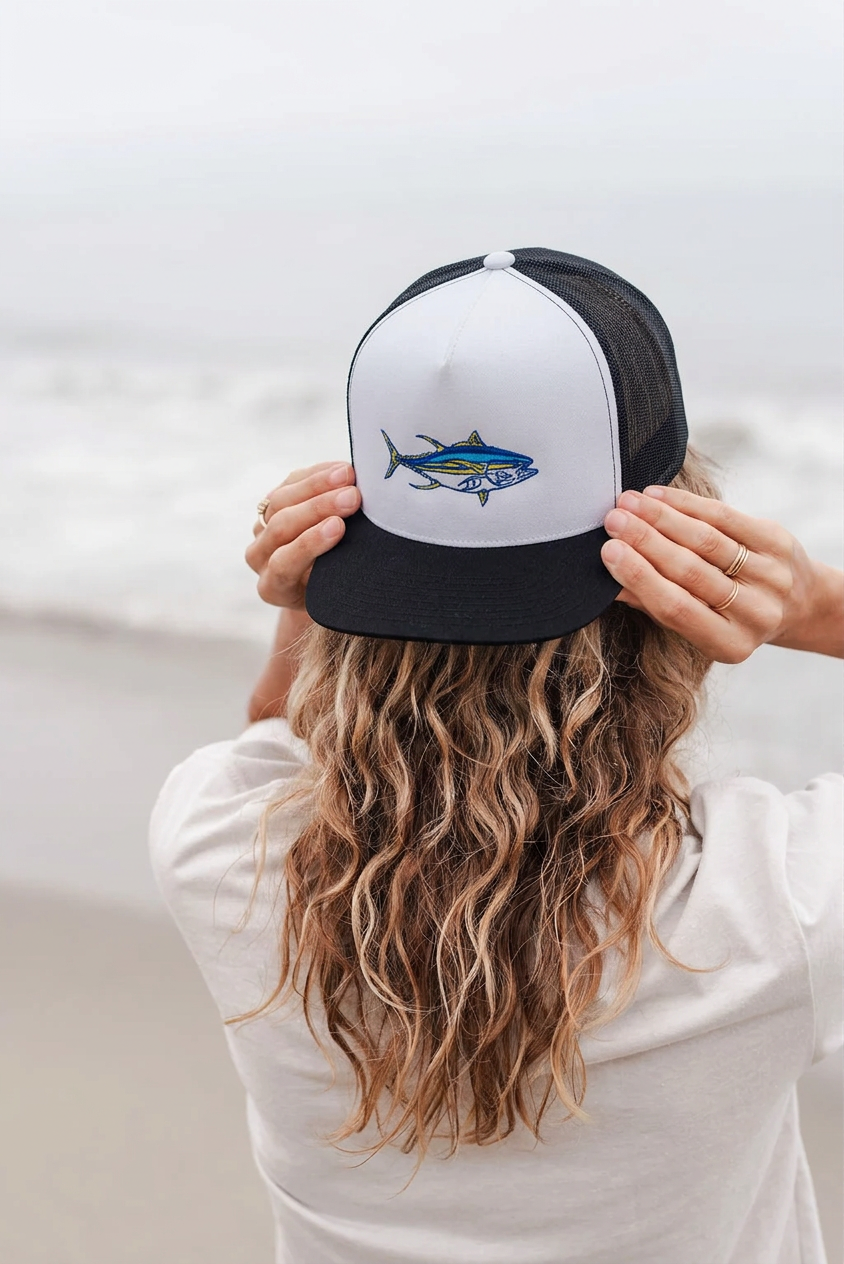 A woman with long wavy blonde hair seen from behind, wearing a black and white trucker hat with a yellowfin tuna graphic on the front, standing on a beach with the ocean in the background.