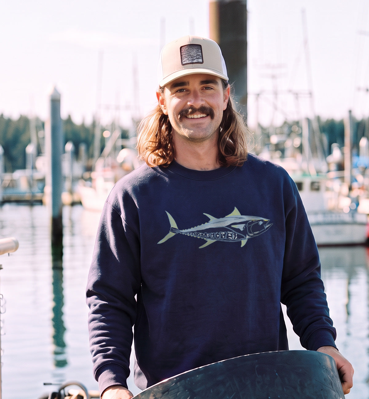 A man with a mustache and long hair stands in a marina wearing a navy blue sweatshirt with a tuna fish graphic and a beige baseball cap.