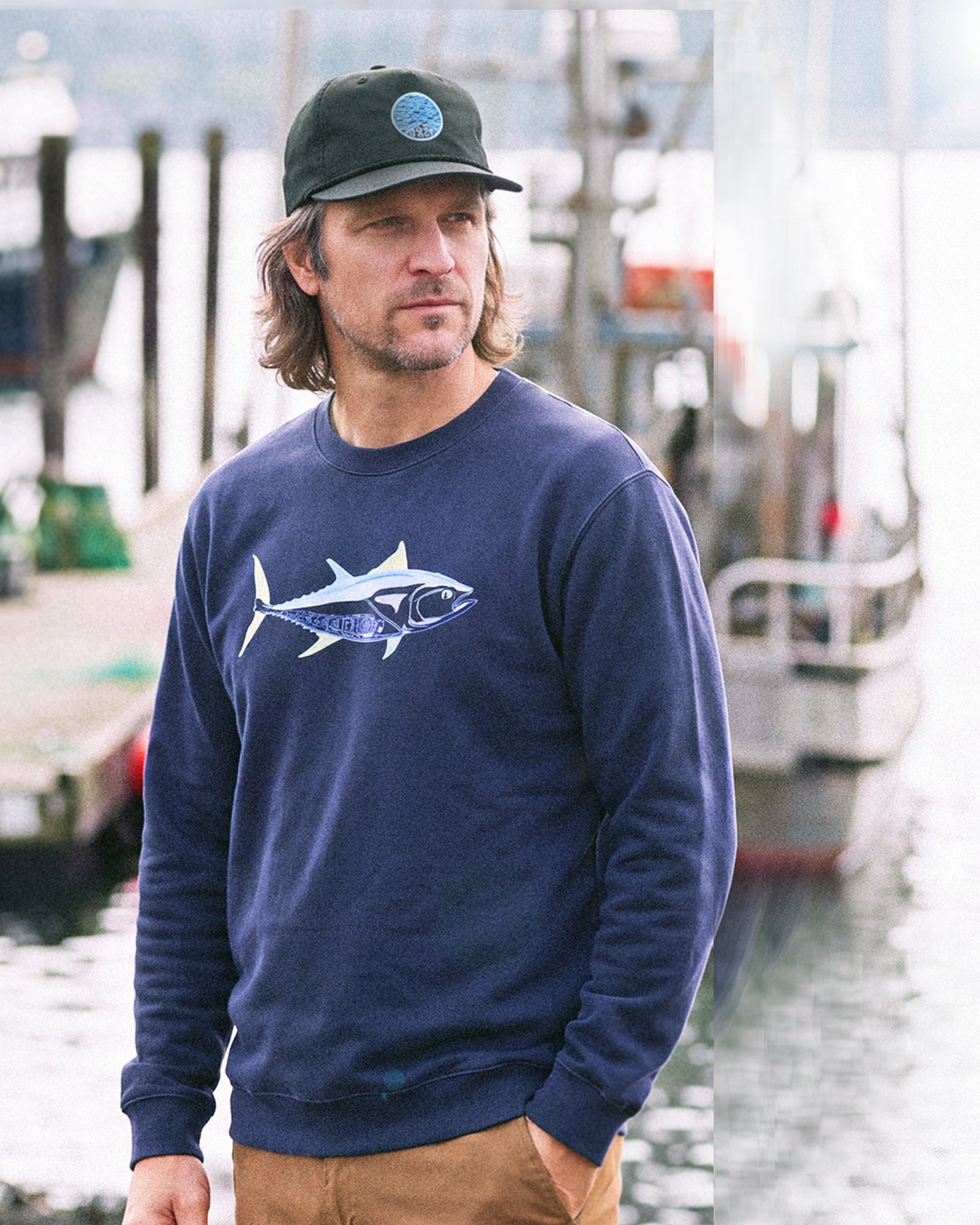 A man wearing a navy blue crewneck sweatshirt with a tuna fish graphic on the chest, paired with a black baseball cap and tan pants, standing in a harbor setting.