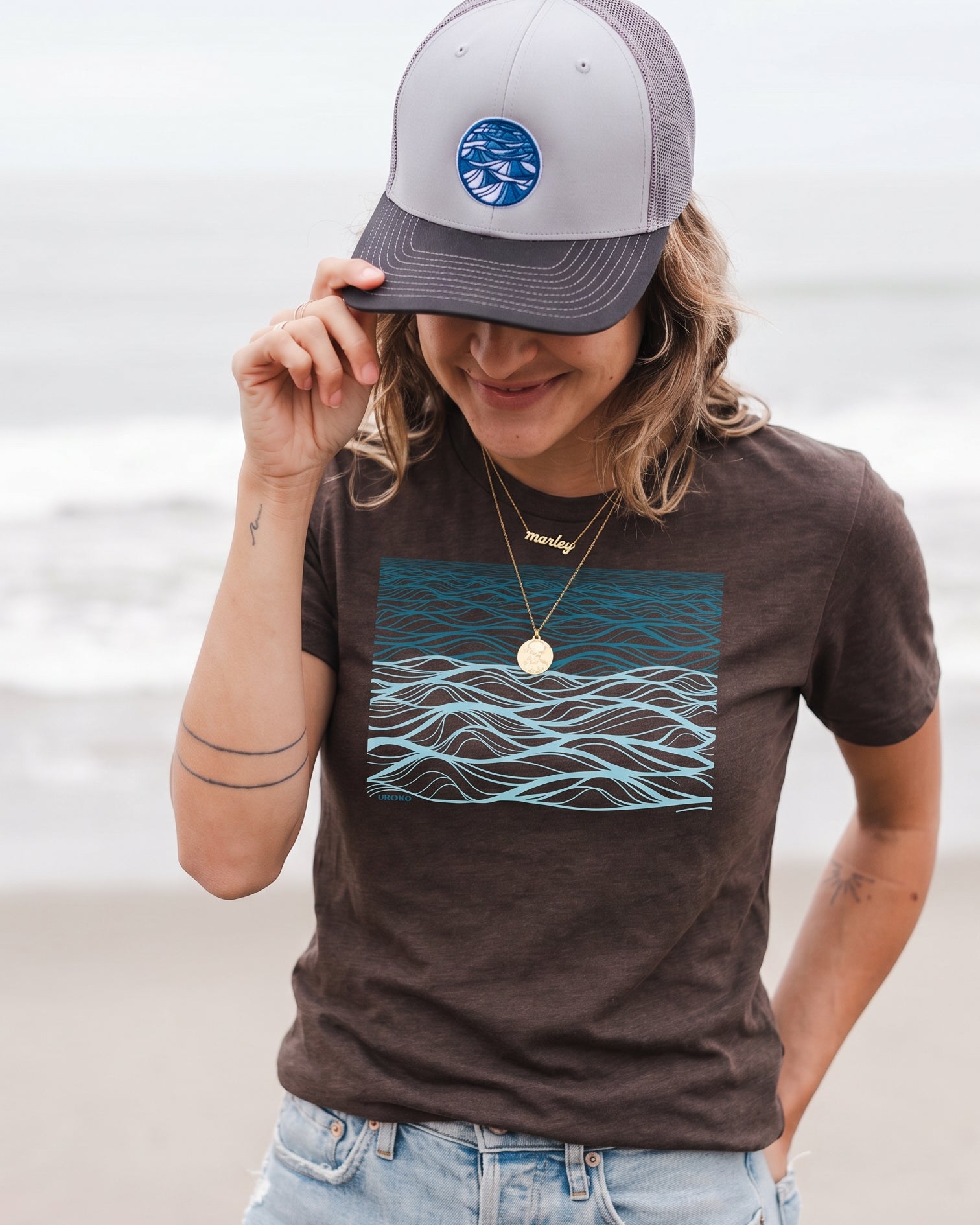 Person wearing a brown t-shirt with a blue wave design and a gray cap on a beach.