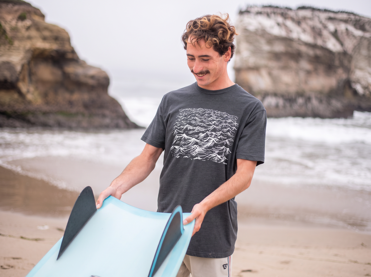 A man standing on a sandy beach holding a light blue bodyboard with black fins. He is wearing a dark gray graphic t-shirt and light-colored shorts, with rocky cliffs and the ocean in the background.