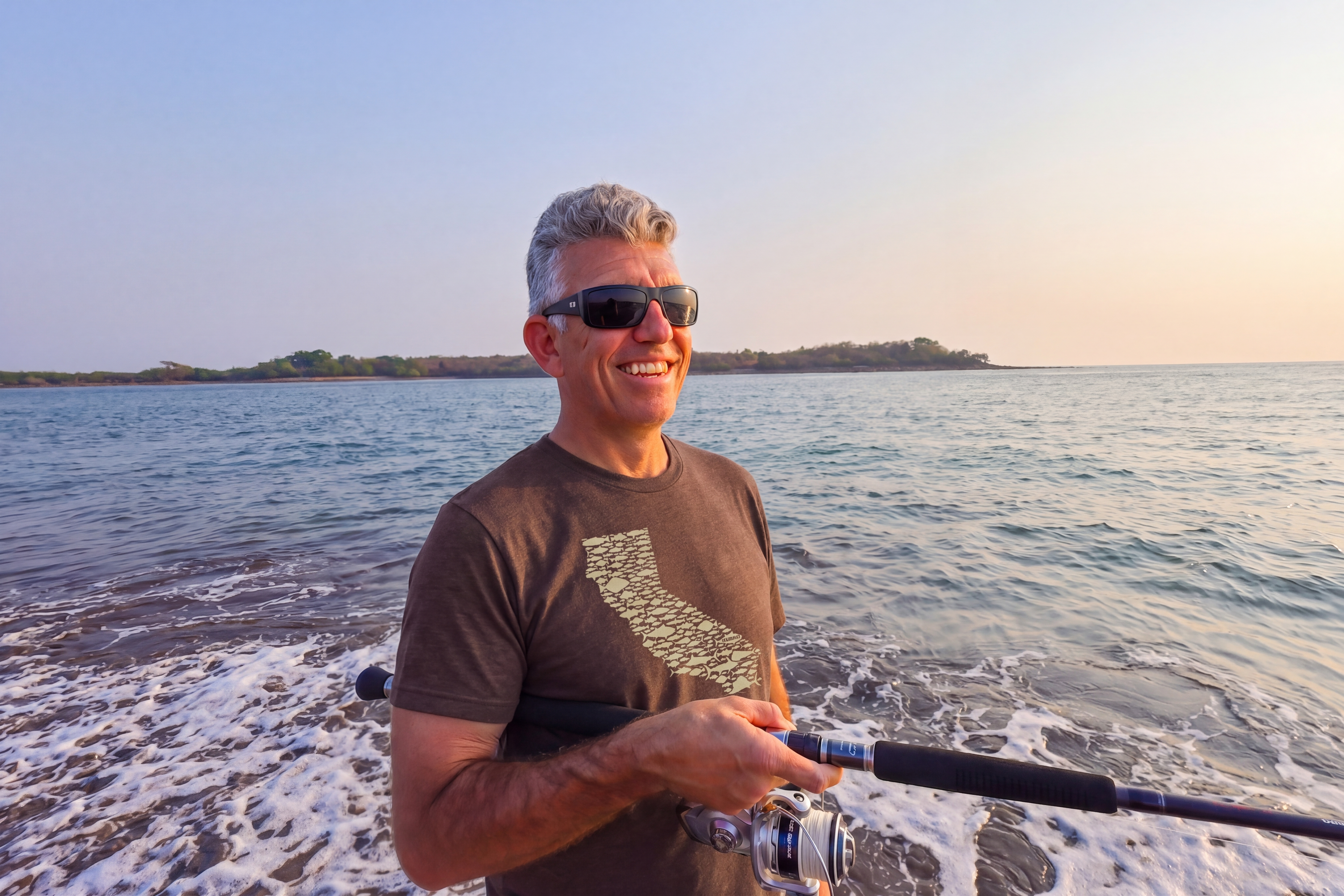 A smiling man with gray hair wearing the California Love fishing tee, holding a fishing rod, with ocean water and distant land under a clear sky.