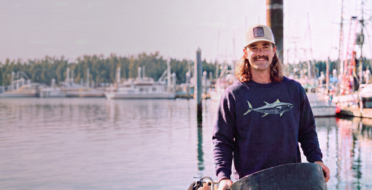 A man with long hair and a beard stands on a dock holding a large black barrel, wearing a navy blue long-sleeve shirt with a shark graphic and the brand name 'Southern Tide' visible, and a beige cap. The background shows a calm harbor with boats and trees under a bright sky.