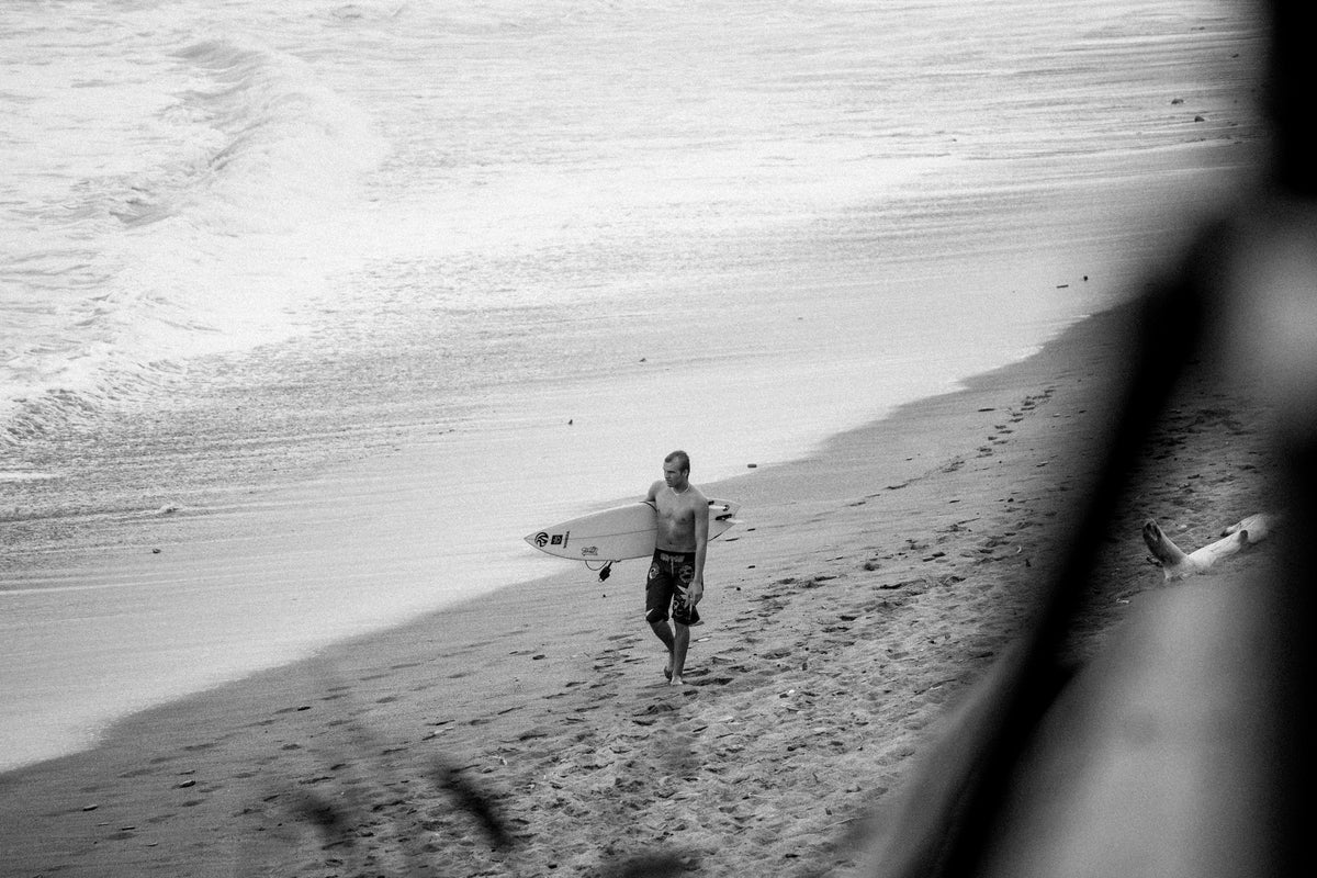 Black and white photograph of Harper King walking on a sandy beach carrying a surfboard.