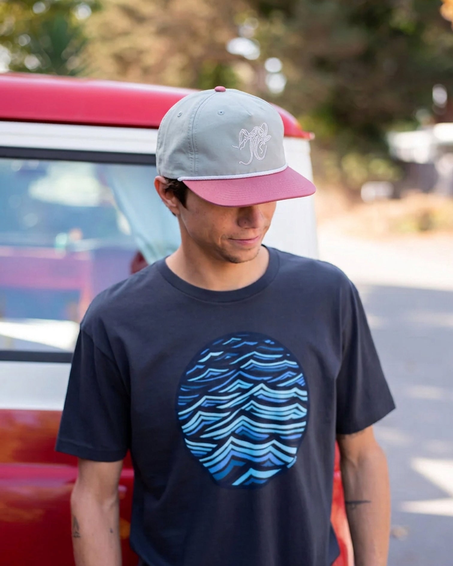 A man wearing a navy blue t-shirt with a blue circular wave graphic on the front, a pink and gray baseball cap, standing in front of a red vehicle outdoors.