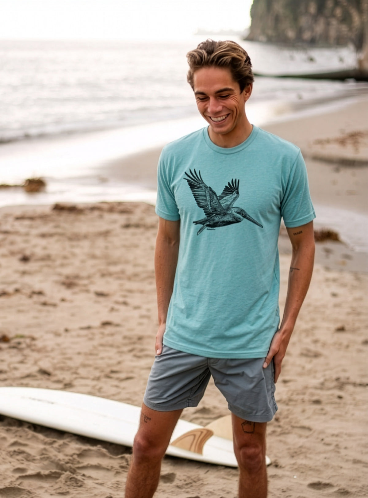 A man standing on a sandy beach wearing a light blue-green t-shirt with a pelican graphic on the chest, paired with gray shorts.