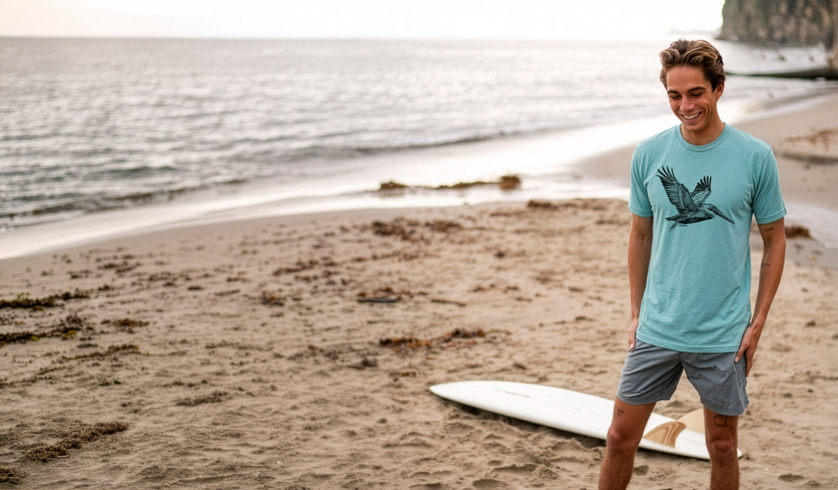 A man standing on a sandy beach wearing a light blue t-shirt with a pelican  graphic and gray shorts, with a white surfboard on the sand and the ocean in the background.