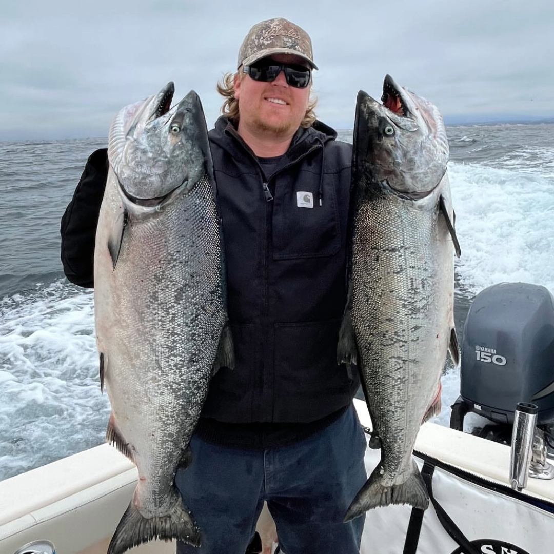 Man holding two large fish on a boat with ocean in the background
