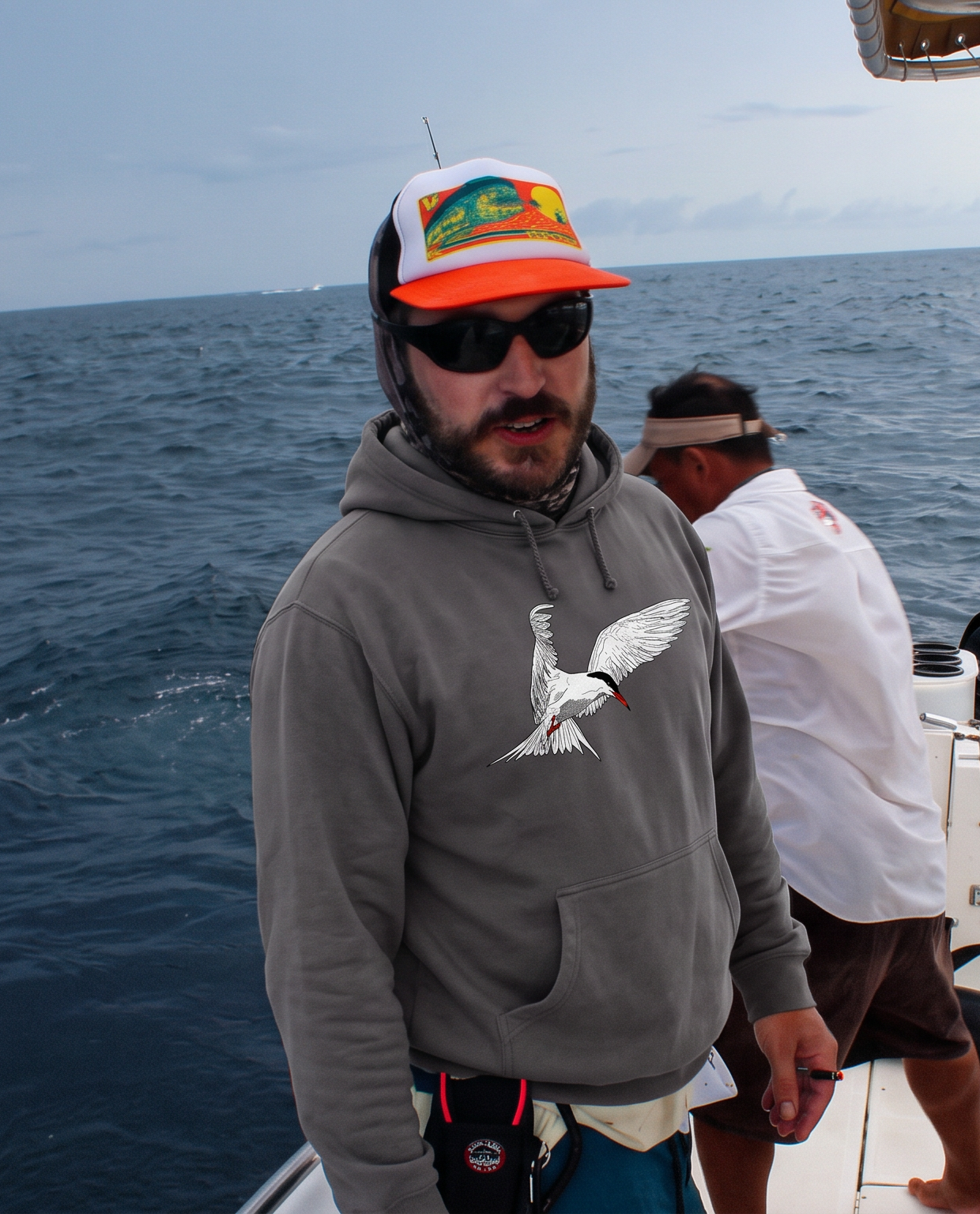 A man on a boat wearing a gray tern hoodie, a white and orange cap, and sunglasses, with another man in the background.
