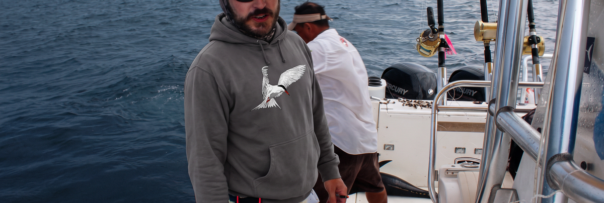 A man wearing a gray hoodie with a tern graphic stands on a boat deck, with ocean water and another person in the background.